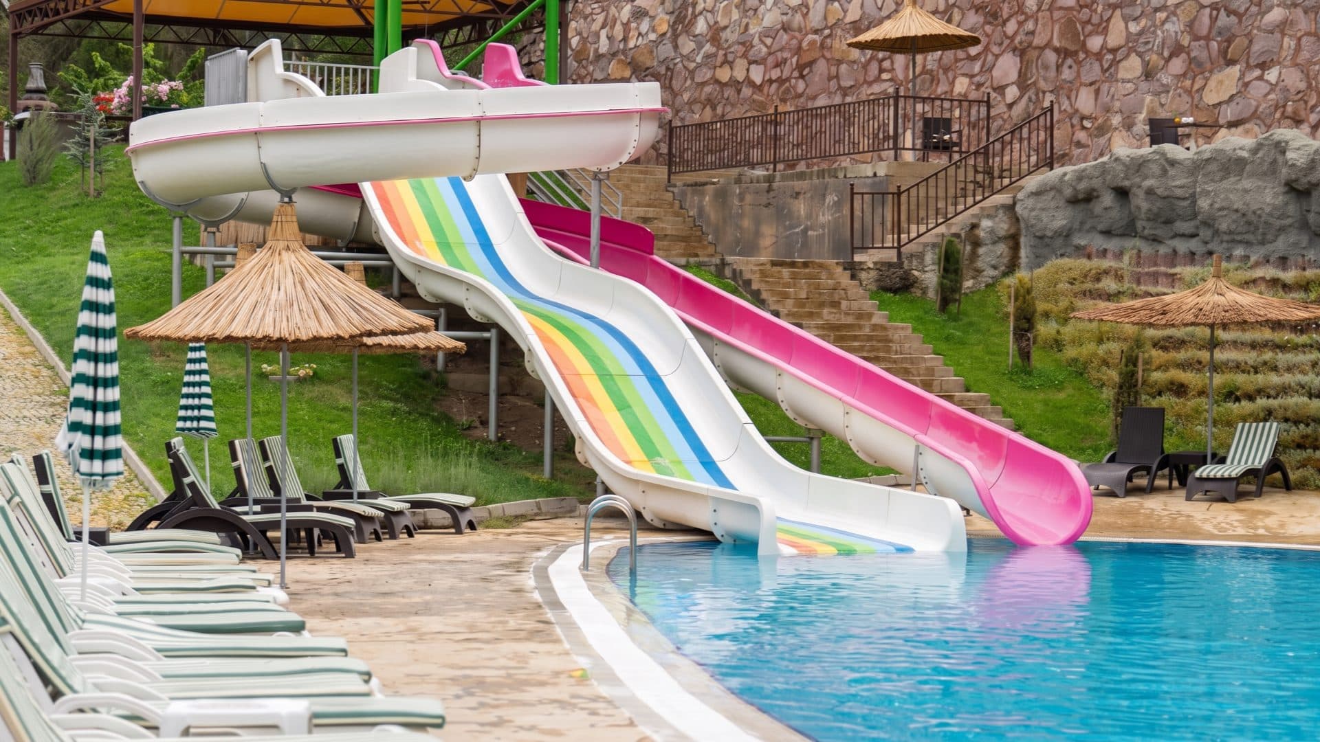Rainbow-striped and pink water slides curving down to the pool edge, with straw parasols and striped loungers alongside.