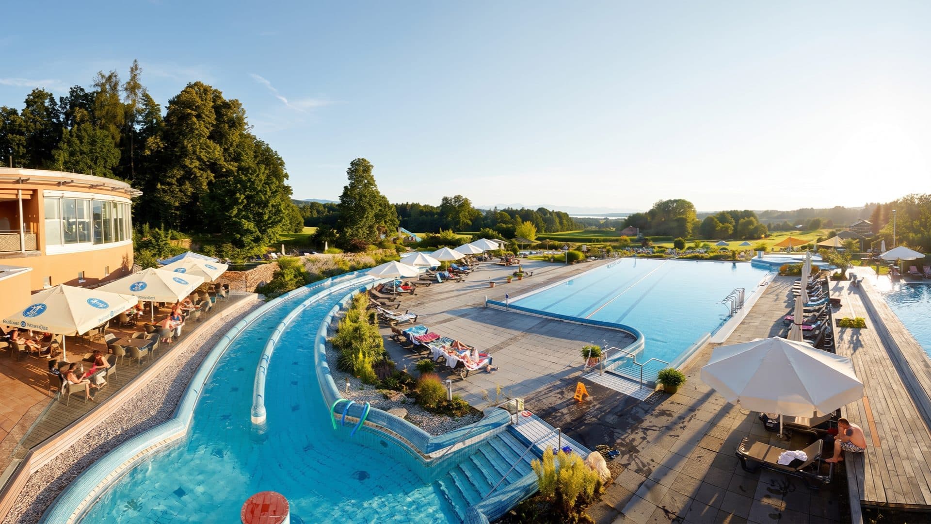 Evening view across the outdoor pools and a curved current channel, loungers under white parasols, with rolling countryside and distant mountains beyond.