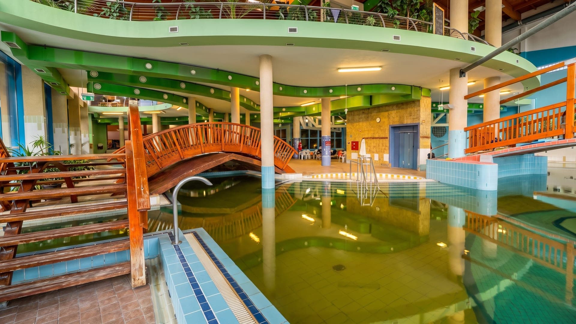 The indoor hall at Cserkeszolo, its green thermal water crossed by a timber bridge under a green-painted gallery.