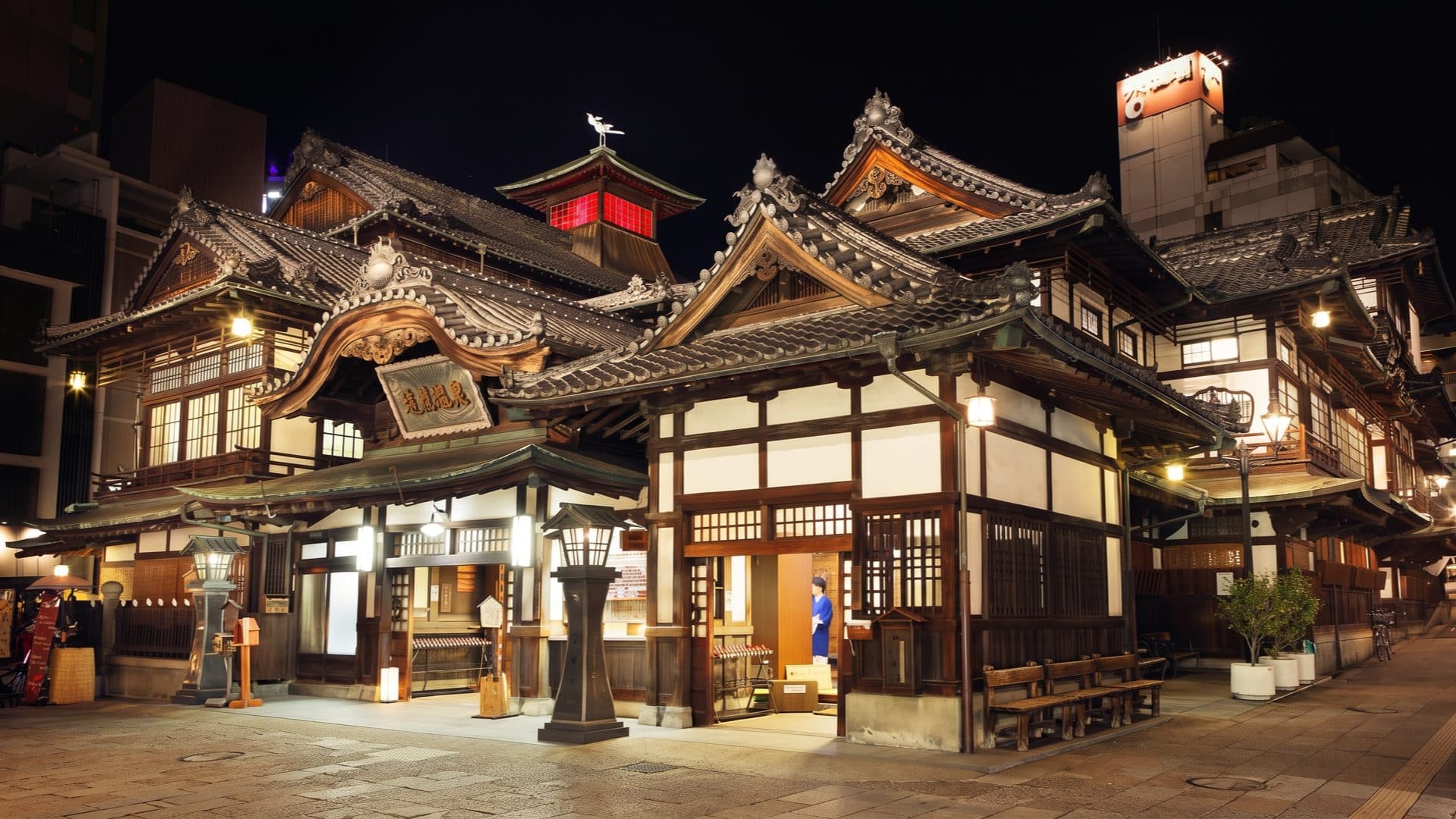 Night view of the Dogo Onsen bathhouse and its ornate tiled roofs, stone lanterns glowing on the plaza and the red-lit turret above.