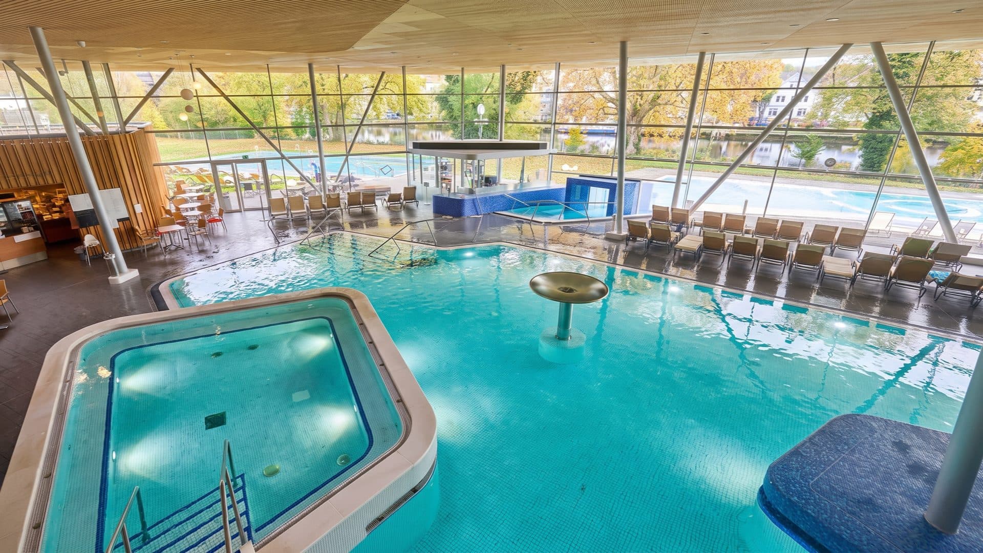 Glass-walled indoor thermal hall with a raised whirlpool and a poolside bar, loungers along a full-height window looking over the river to the outdoor pools.