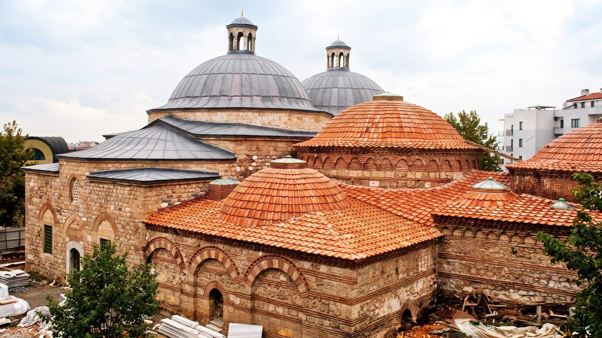 A cluster of tiled and lead-covered domes with lantern cupolas on the roof of a historic Ottoman bathhouse, arched brick walls below.