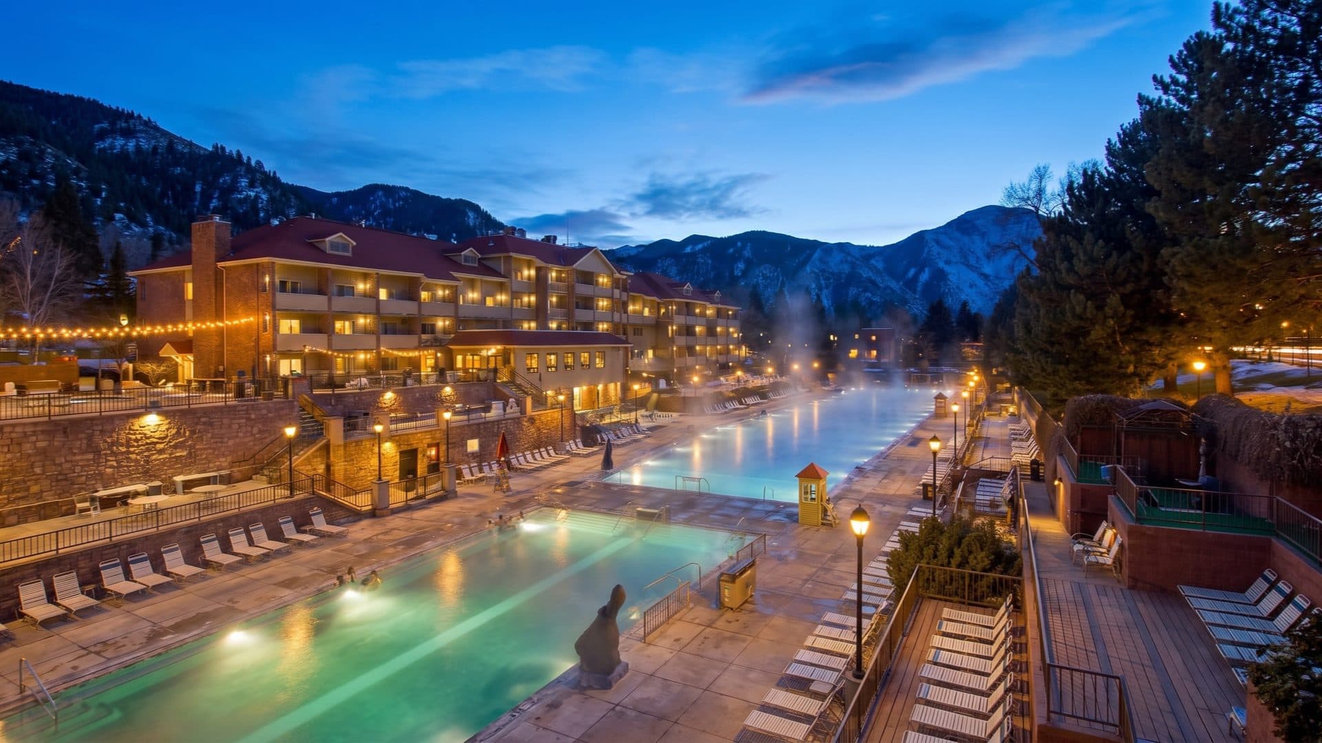 Evening view of the resort pools steaming under strings of lights, a lit lodge and snow-dusted mountains beyond.