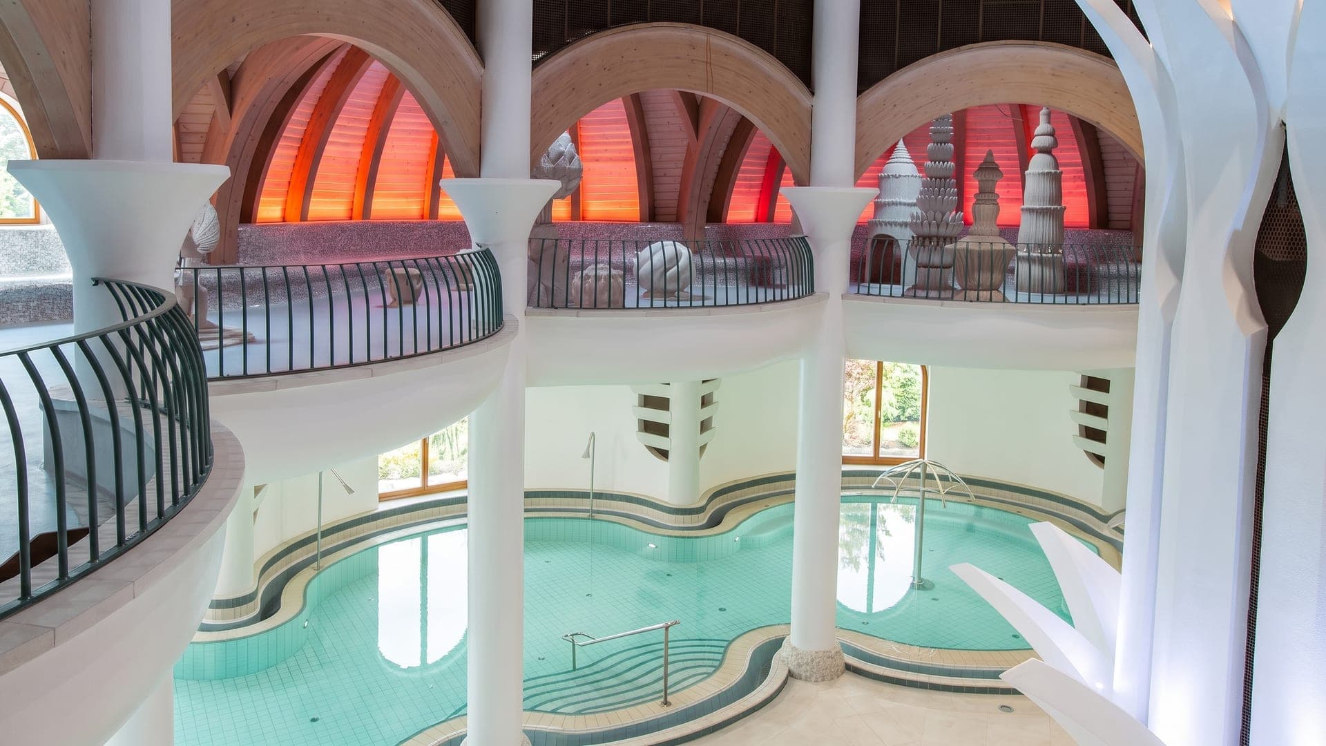 Curving indoor pools beneath white columns and red-lit vaulted arches