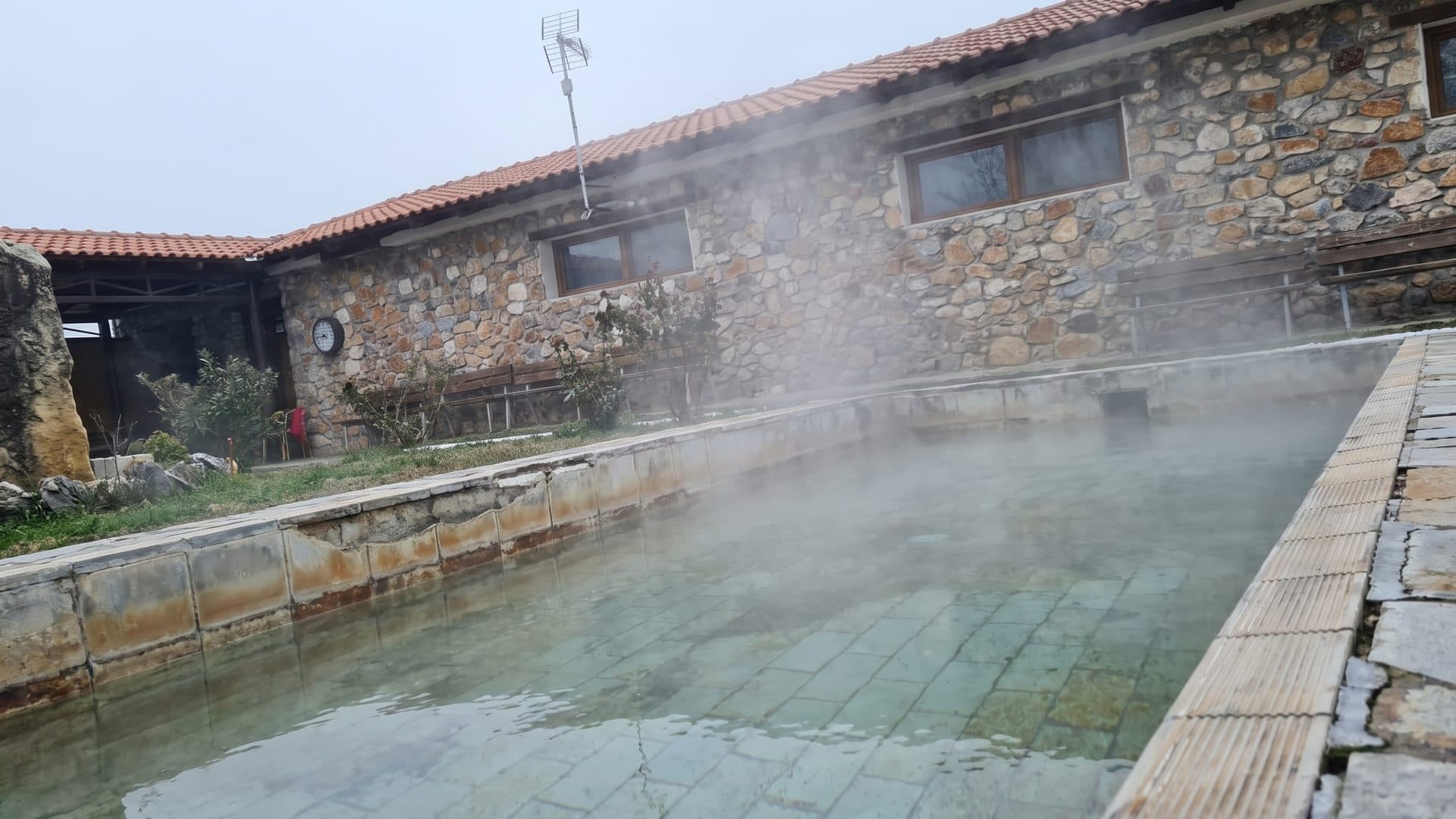 Steaming open-air mud pools in front of a long stone building with a timber-framed upper floor.