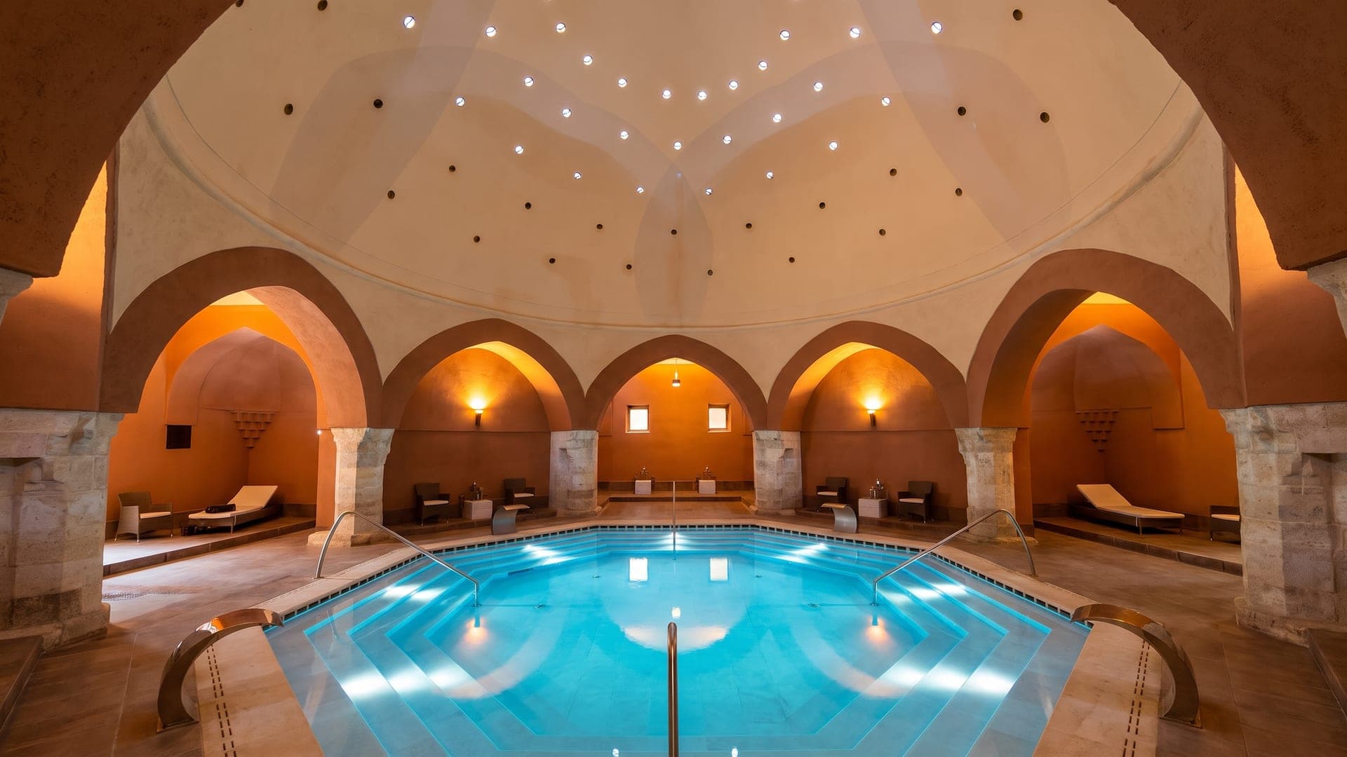 The dome of the Veli Bej bath in Budapest seen from below, daylight falling through the star-shaped openings onto the water.