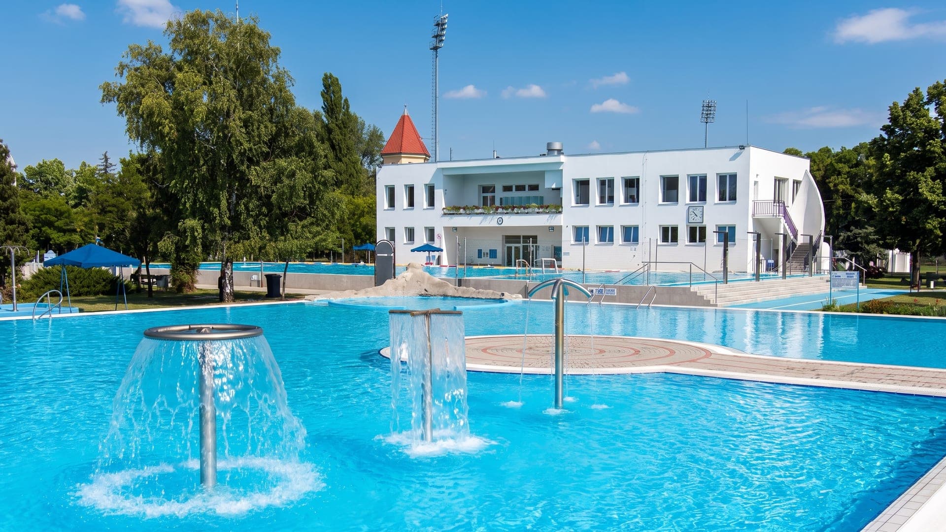 The outdoor pool at the Kecskemet bath, with mushroom fountains and the white bath building behind.