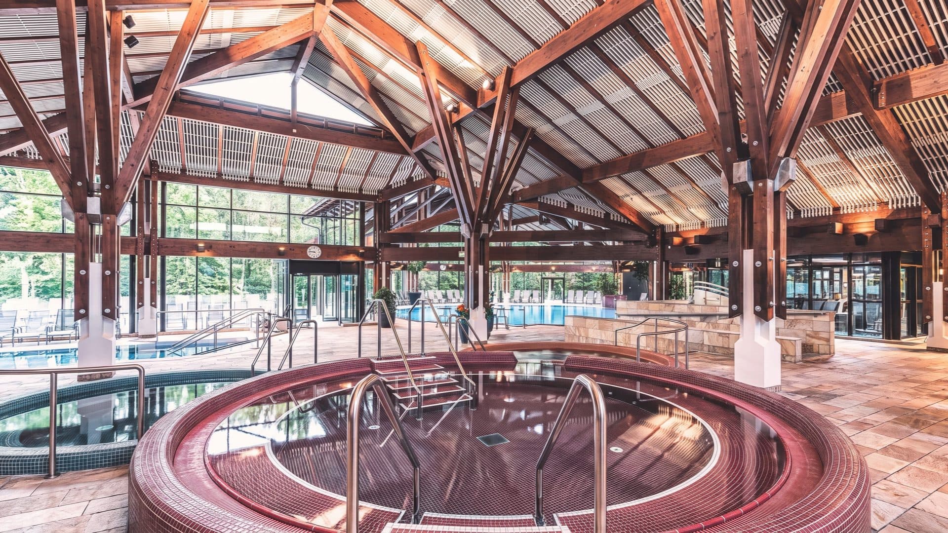 Indoor thermal hall under a grand exposed-timber roof, a round red-tiled whirlpool in front of the main pool and glass walls opening to the forest.