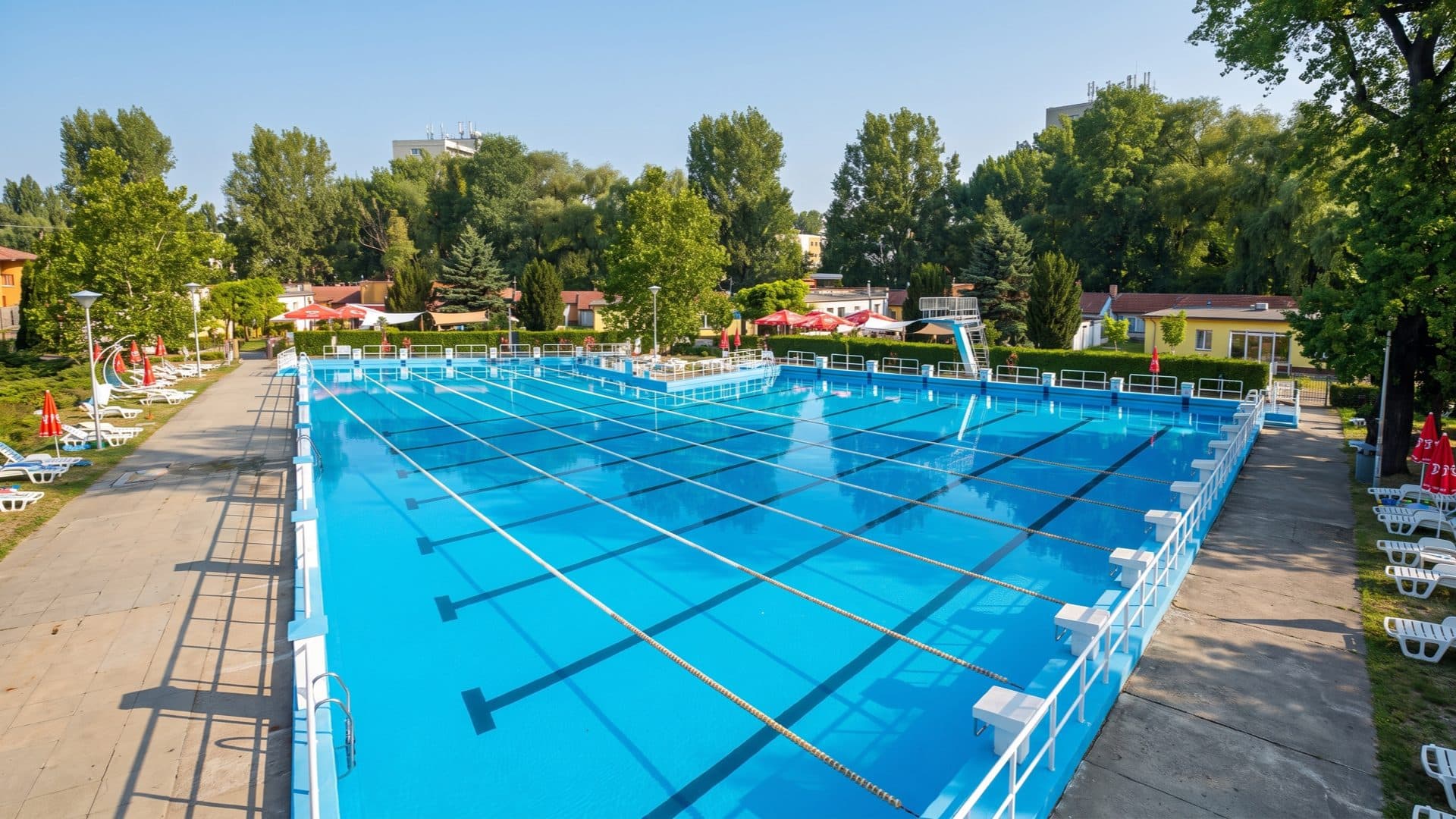 Large outdoor swimming pool marked with lanes and starting blocks, poplars and red parasols around the deck.