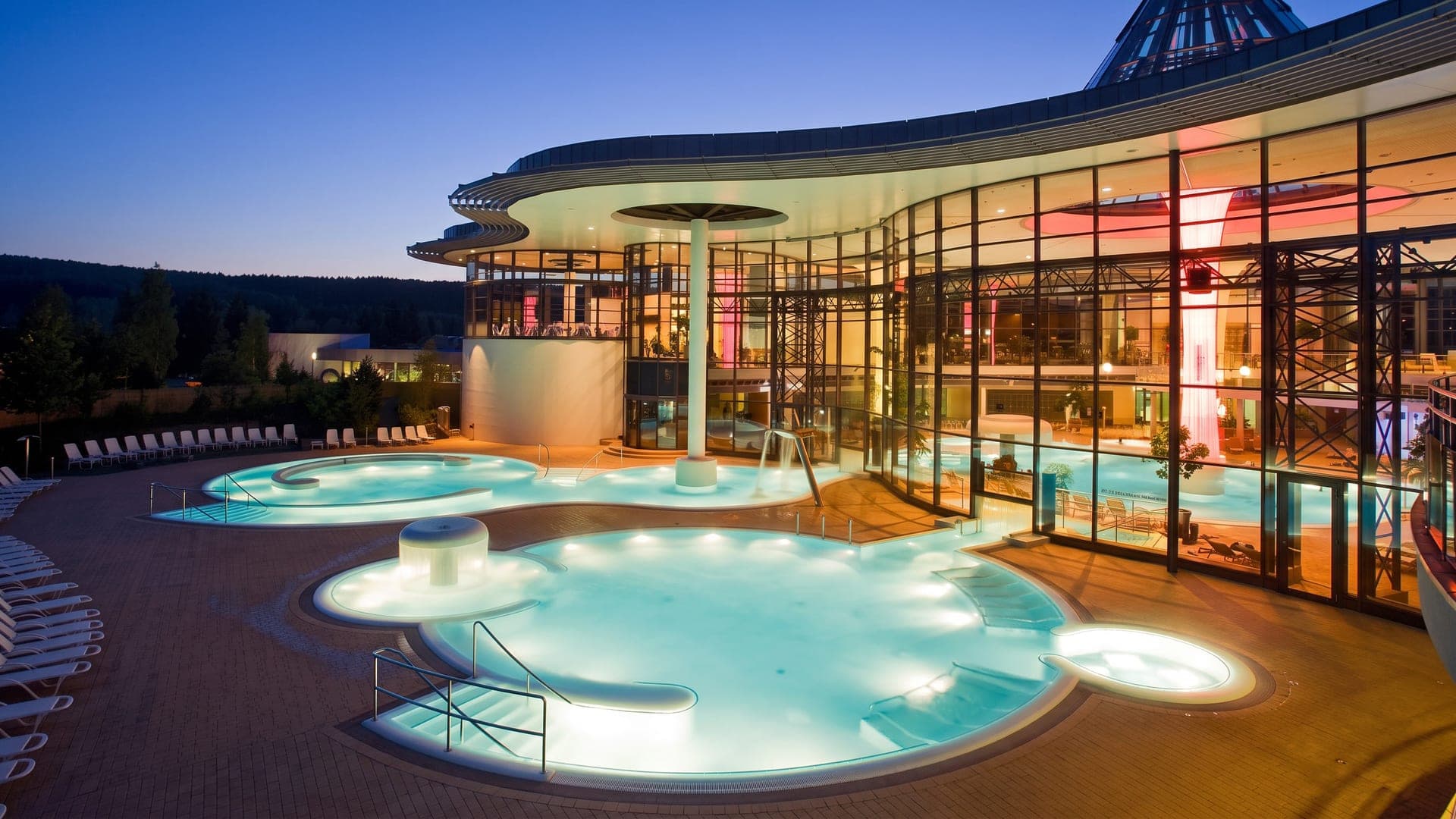 The curved glass building at dusk, its outdoor pools lit from within the water