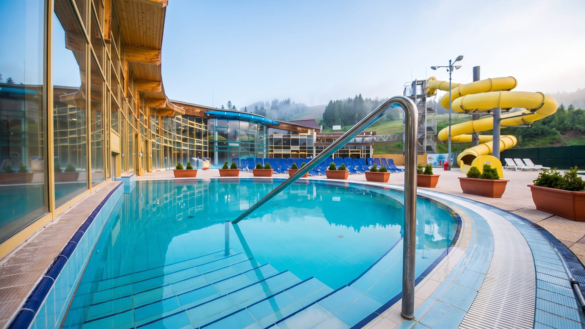 The outdoor pool at the Oravice baths, with a yellow waterslide and the wooded hillside beyond.