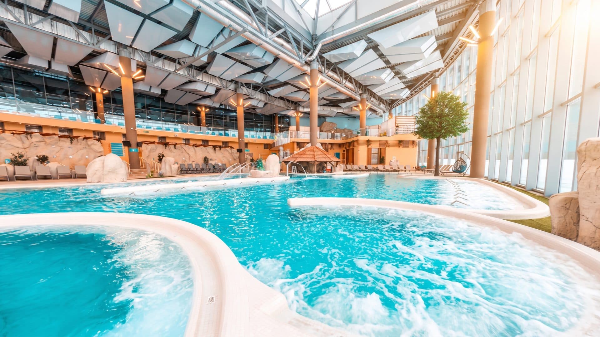 Bubbling indoor thermal pools under a high steel-and-glass roof, with faux-rock features in a bright, airy hall.