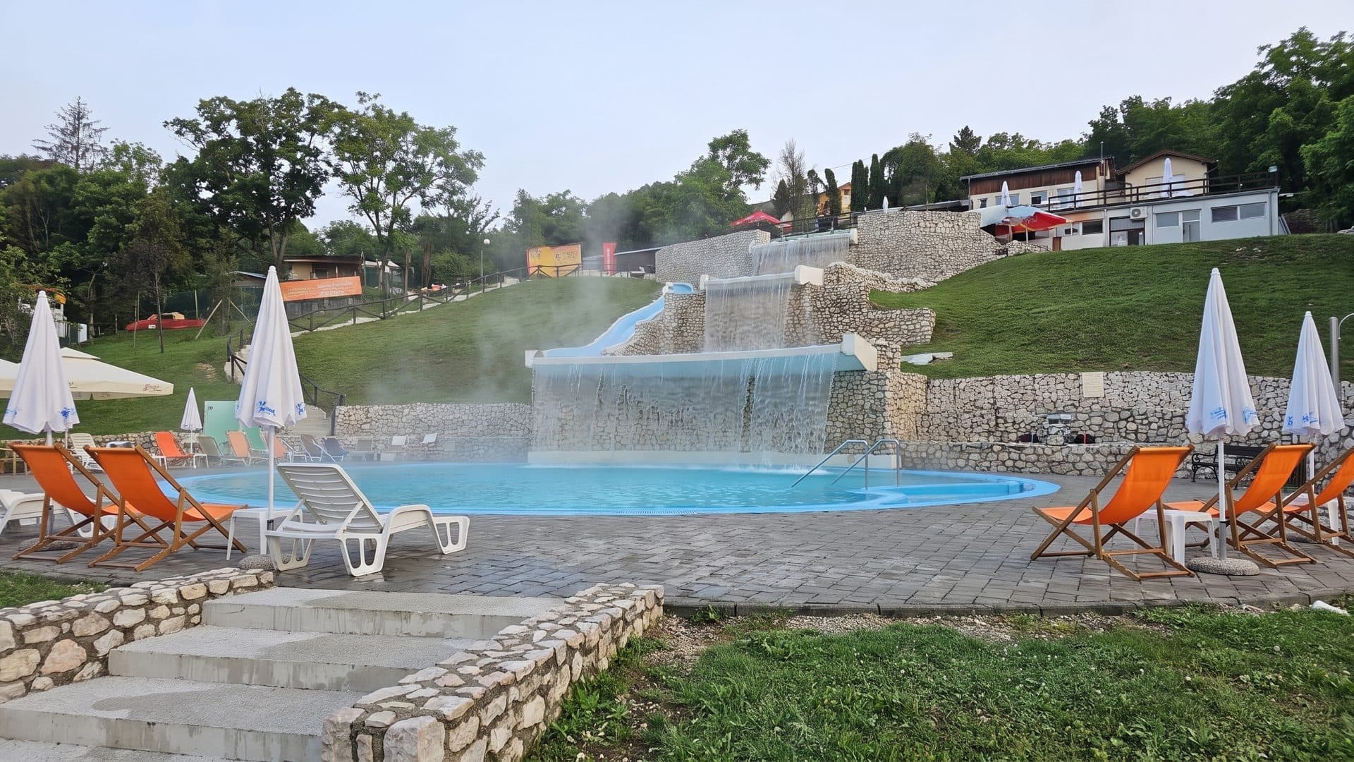A thermal pool at Panonska jezera fed by a wide waterfall over a stone wall, orange deckchairs on the deck and steam over the water