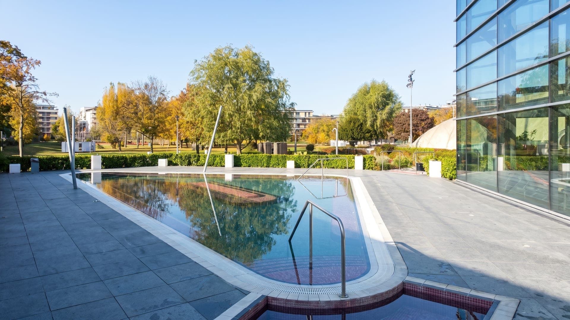 The outdoor pool at the Paskal bath in Budapest, its long basin beside the glass hall with autumn trees behind.