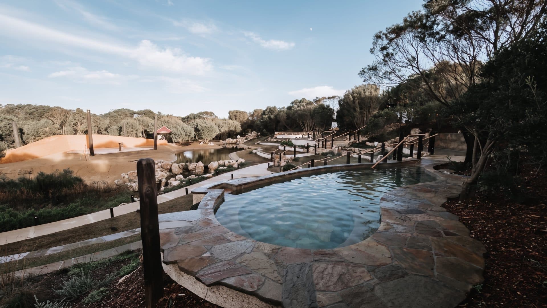 A round flagstone pool on terraced spa grounds, with boardwalks, a sunken amphitheatre and native bush stretching down the slope.