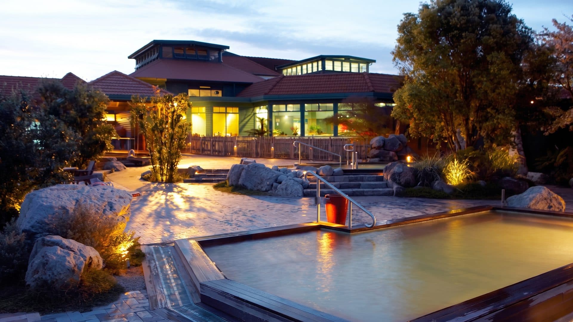 The lakeside spa pavilions lit at dusk, with landscaped rock pools and boulders in the foreground.