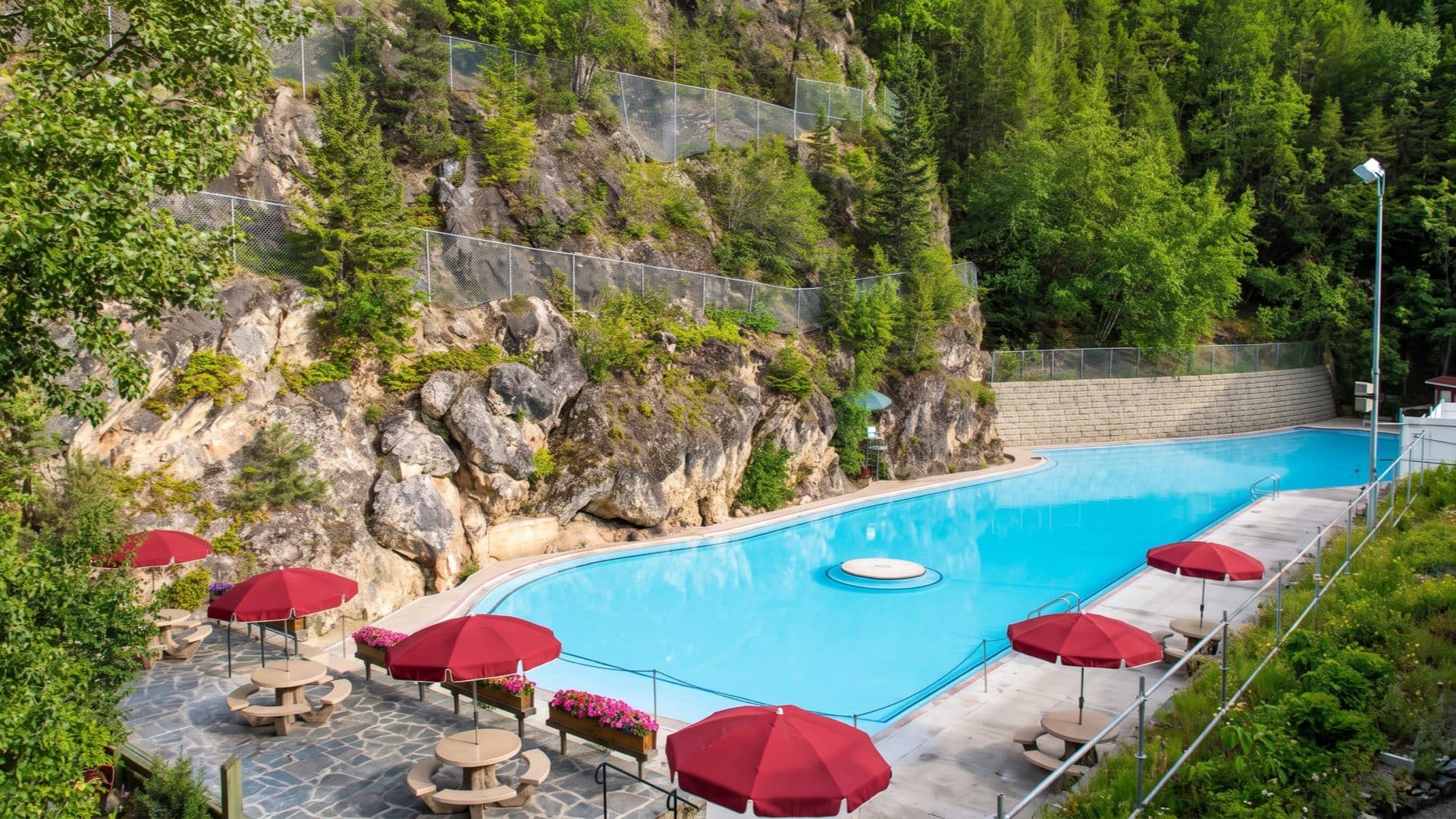 The canyon hot-springs pool in summer, red parasols along the deck beneath green forested cliffs and rock outcrops.