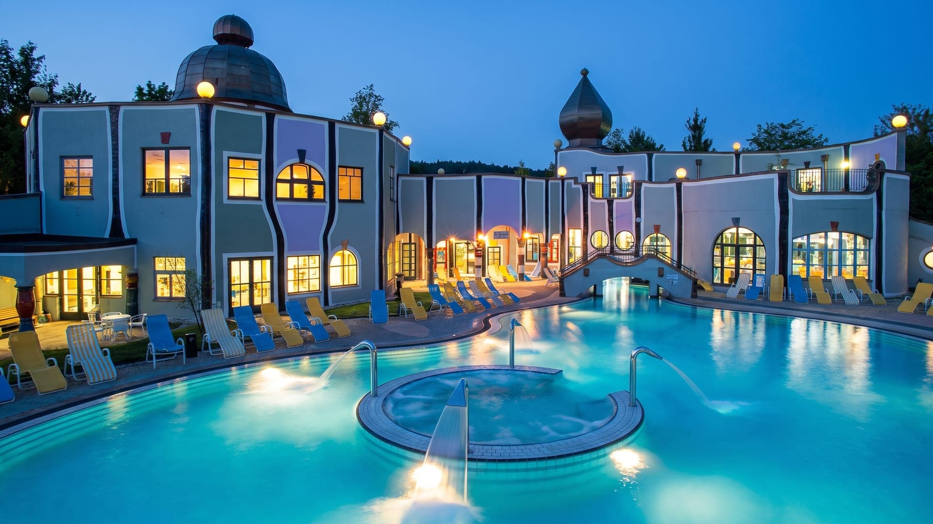 The blue facades of Rogner Bad Blumau lit at dusk, reflected in the large outdoor thermal pool.