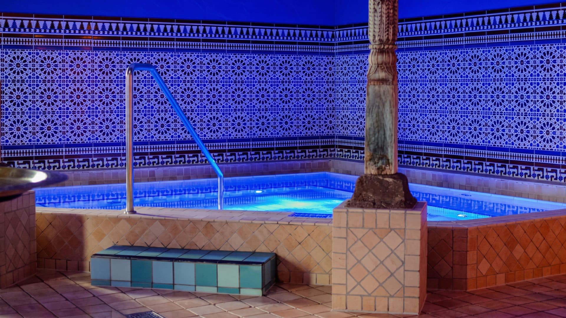 Whirlpool set against walls of intricate blue Moroccan tilework, with a carved column and a warm terracotta floor.