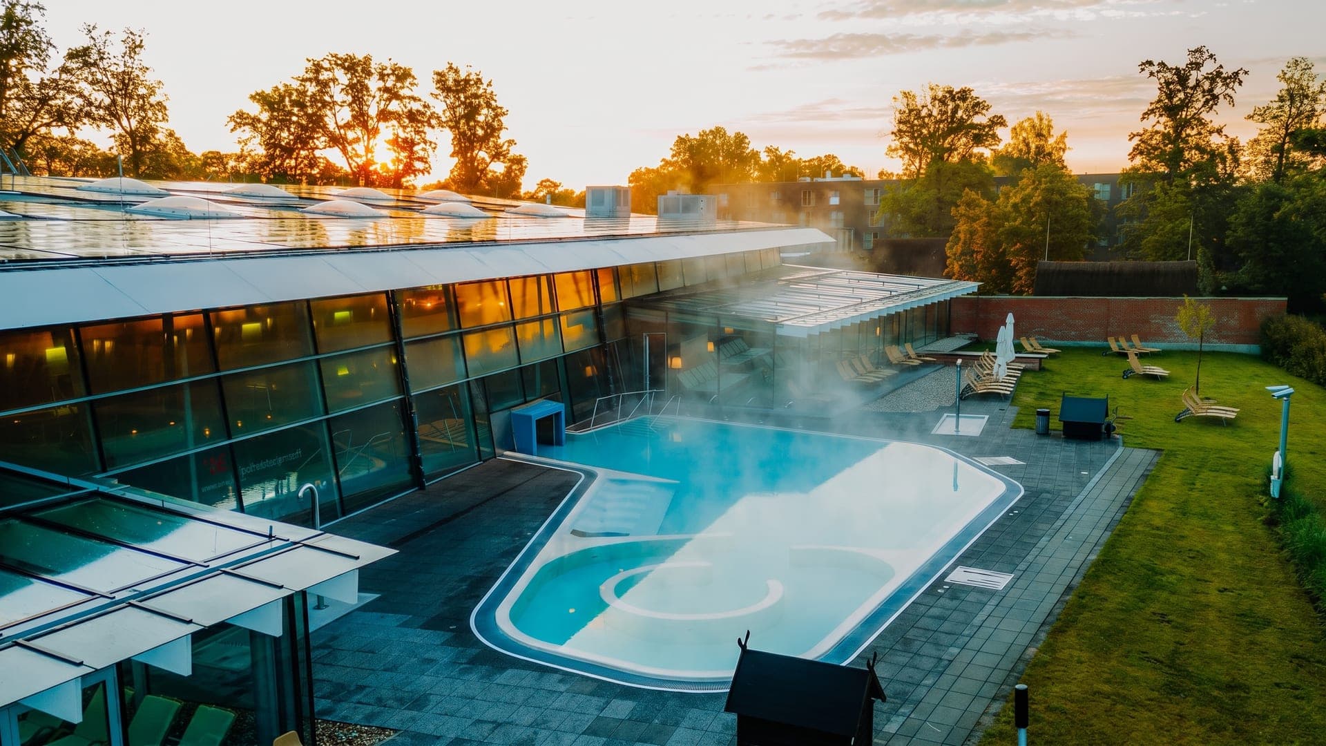 The outdoor pool at the Spreewald Therme in Burg seen from above, its curved basin beside the glazed hall among trees.