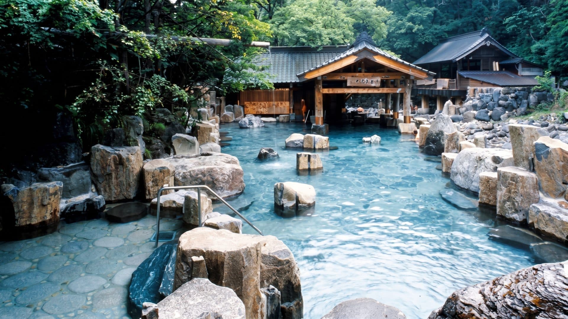 Large open-air rock bath among boulders, a timber bathhouse pavilion behind and forested hillside all around.