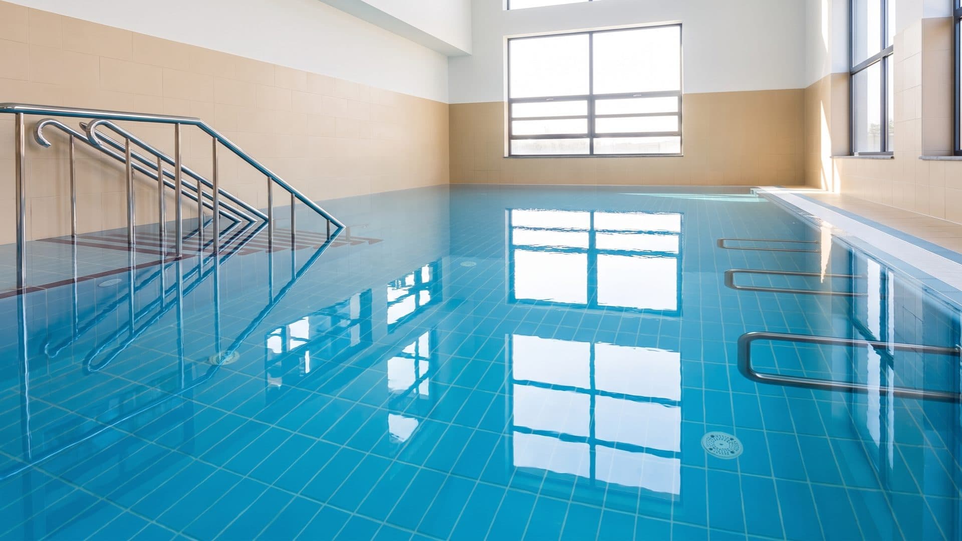 Plain indoor therapy pool tiled in blue, with stainless handrails and tall bright windows.