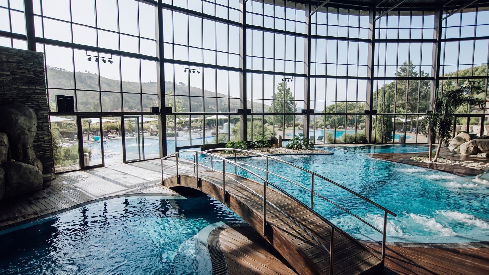 Curved indoor thermal pools joined by a timber footbridge, a full-height glass wall opening to outdoor pools and hills.