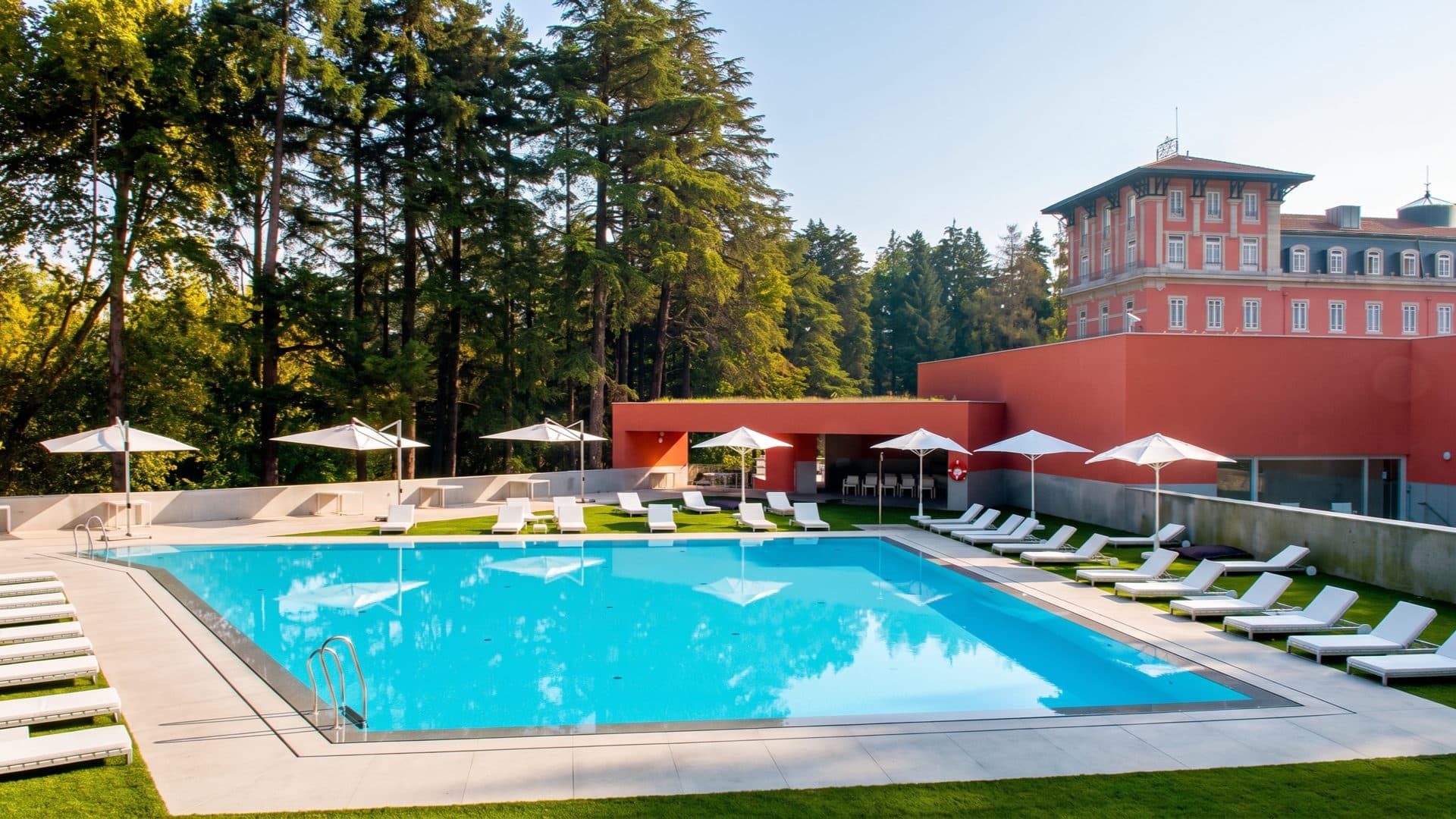 Outdoor pool with white loungers and parasols, the pink belle-epoque palace hotel rising behind tall pines.