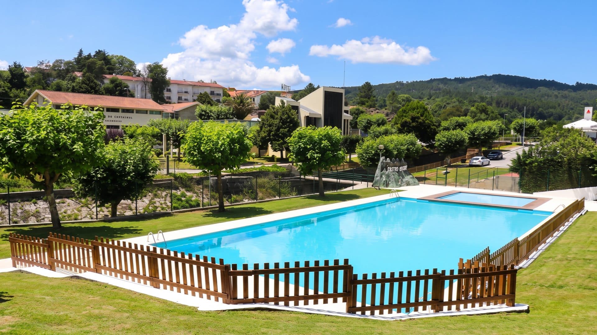 Open-air rectangular pool behind a timber picket fence, lawns and a hillside town beyond under a blue summer sky.