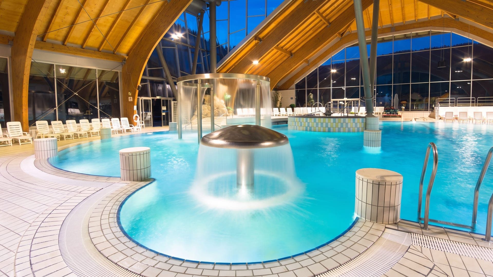 The indoor pool at Terme Snovik under curved timber arches, with a mushroom fountain pouring into the water.