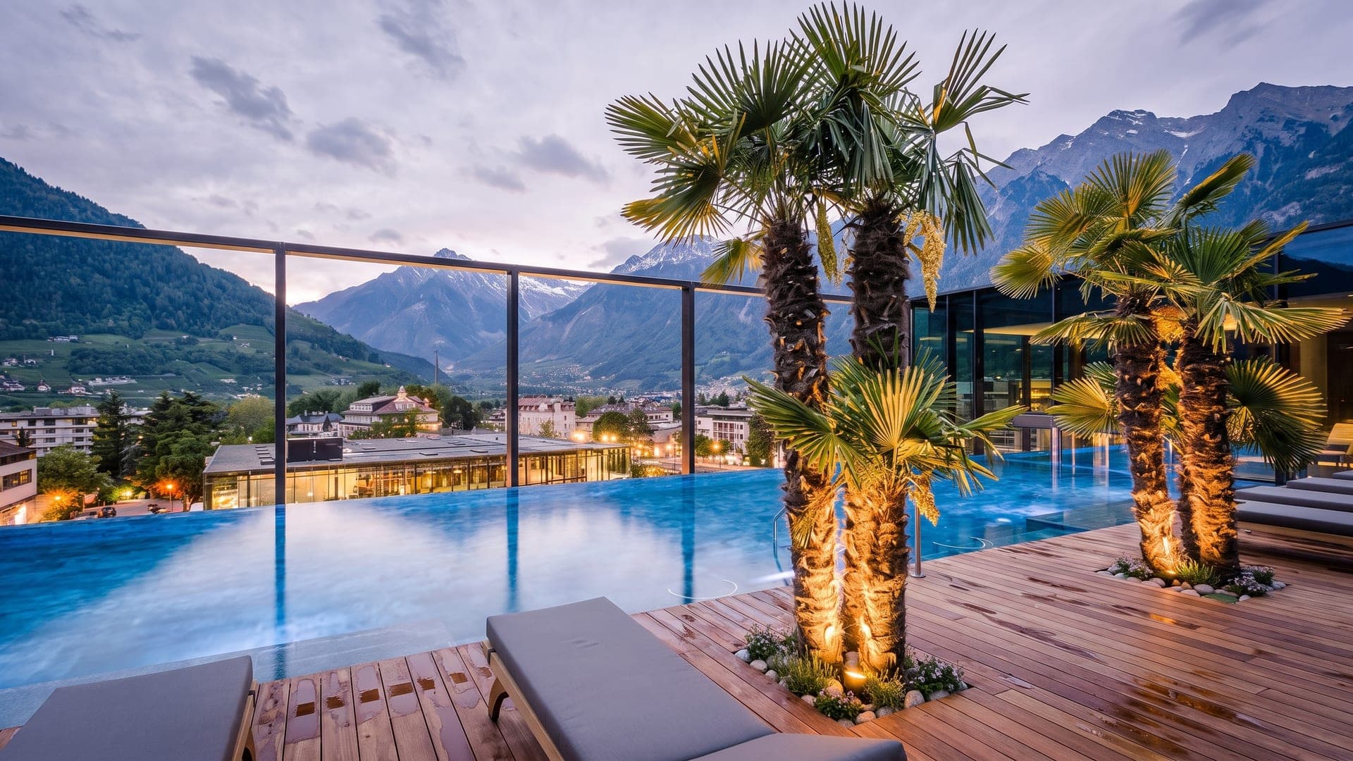 The rooftop pool at the Therme Meran at dusk, palms along its edge and the mountains above the town.