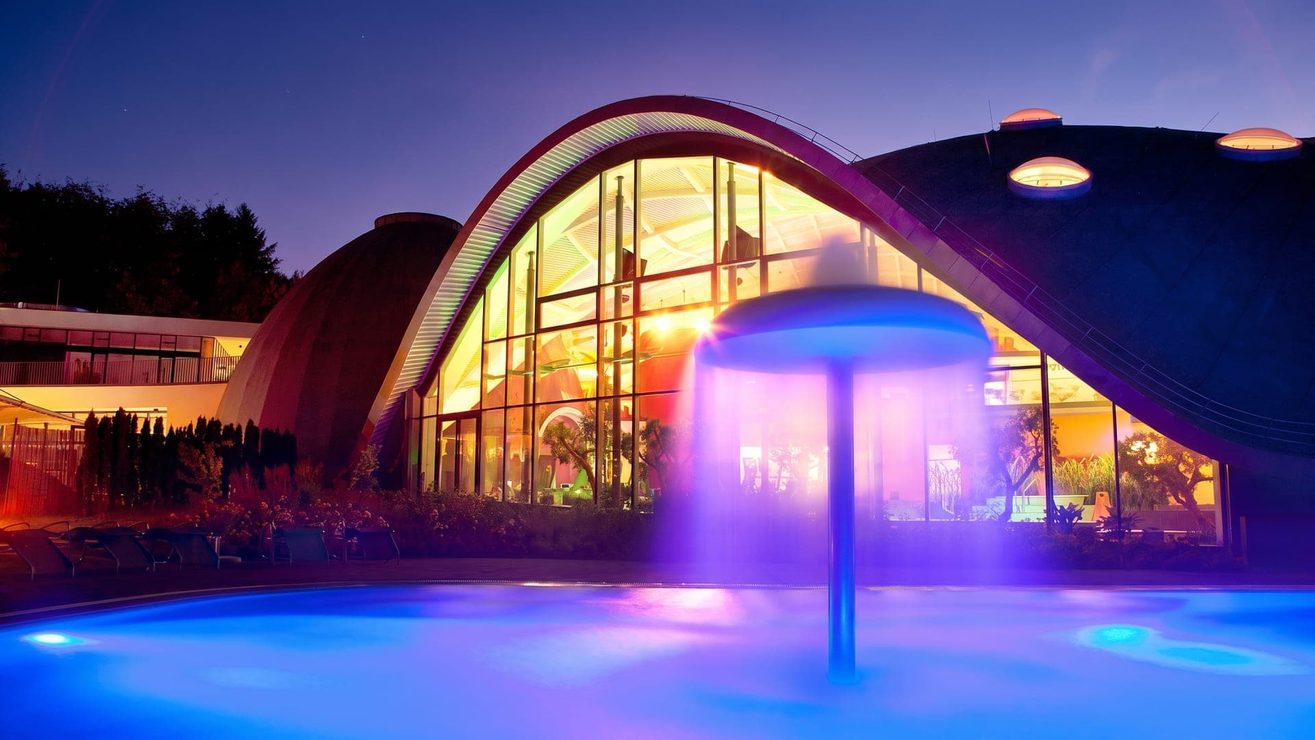 The outdoor pool at the Toskana Therme in Bad Orb at night, a lit mushroom fountain pouring into the water below the wave-shaped roof.