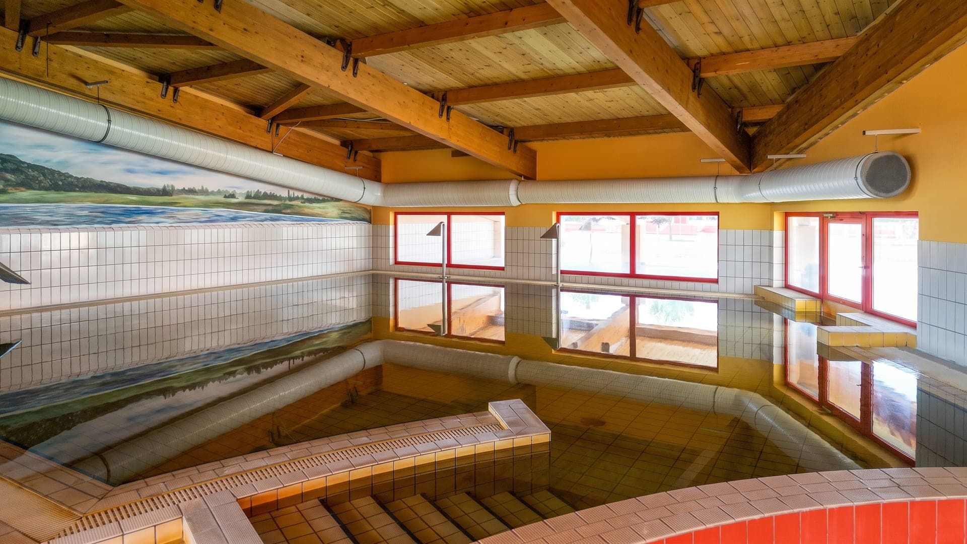 The indoor pool at the Vulkan bath in Celldomolk, its green thermal water under a timber roof with a painted mural along the wall.