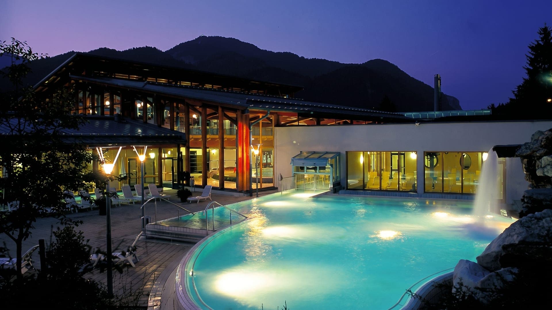 The outdoor pool at the Watzmanntherme in Berchtesgaden at dusk, the mountains in silhouette above the lit water.
