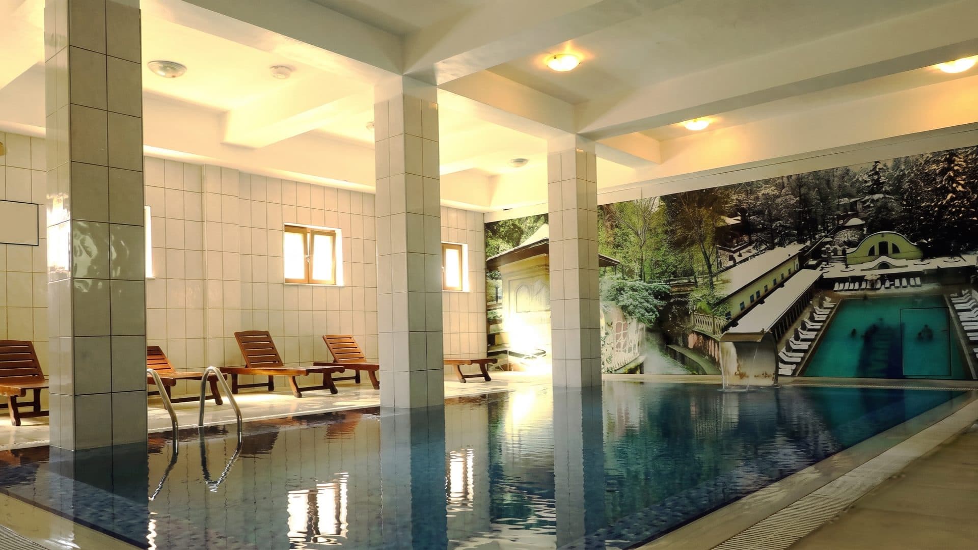 Indoor thermal pool with tiled columns and wooden loungers, a large mural of the historic bath complex along one wall.